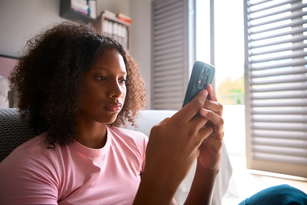 A young woman with curly hair wearing a pink shirt sits indoors, focused on her smartphone. Sunlight streams through partially open blinds in the background, illuminating the modern, cozy room.