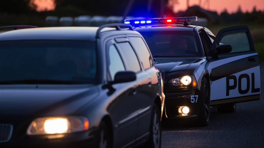 A police car with flashing red and blue lights is parked behind a dark vehicle on the roadside at dusk, with the police car’s driver-side door open.